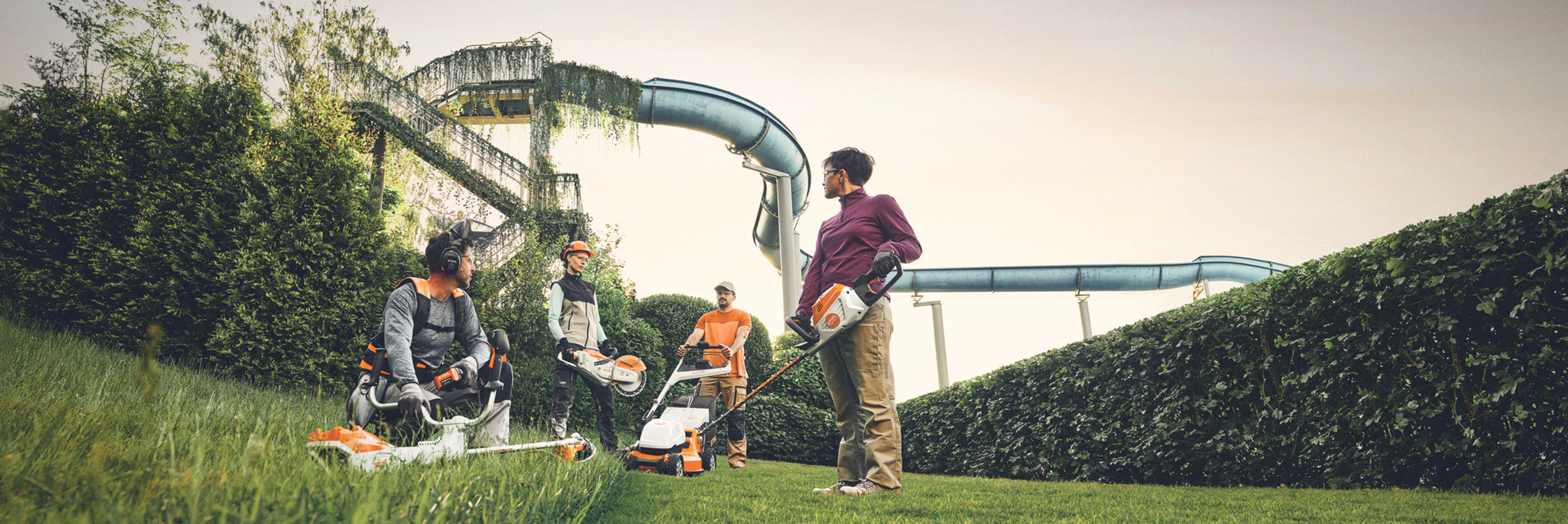 En gruppe mennesker arbejder med STIHL haveredskaber i et grønt område med en stor hæk og vandrutsjebaner i baggrunden. De er udstyret med buskryddere, plæneklippere og beskyttelsesudstyr.