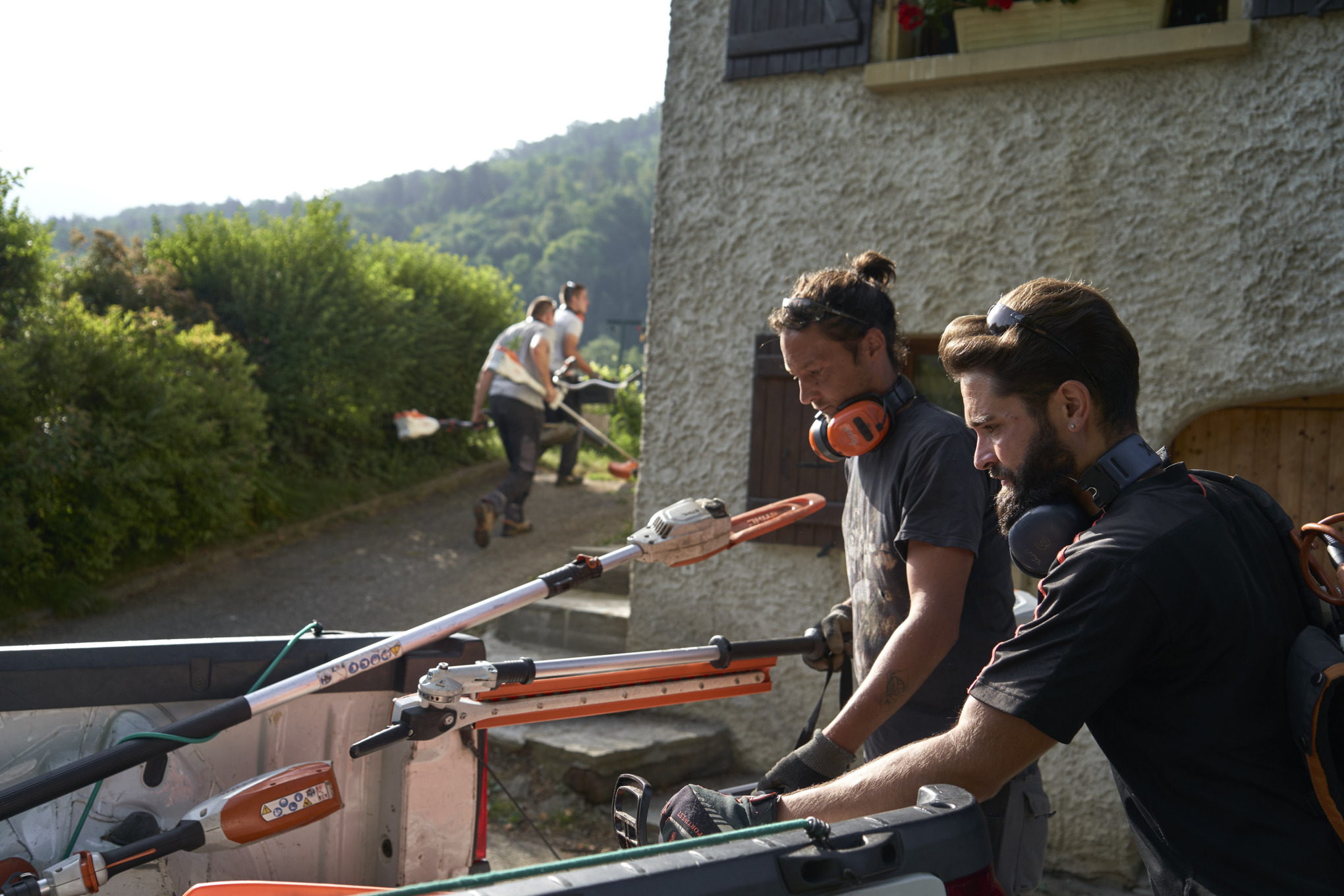 To landskabsgartnere læsser STIHL batteriværktøj af en bil med andre arbejdere i baggrunden 