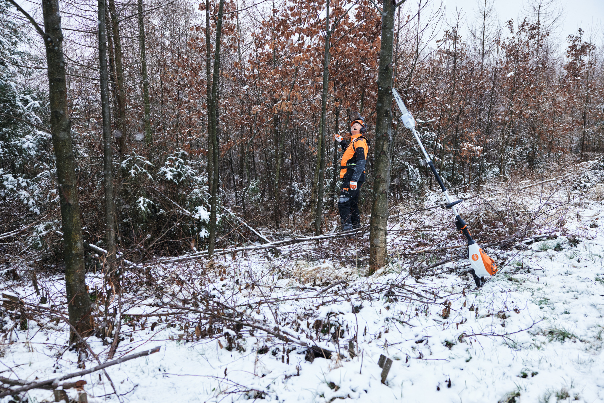 En mand i beskyttelsesudstyr,  der arbejder i en snedækket skov, læner sig op ad en træstamme i forgrunden med en STIHL HTA 150 batteridrevet stangsav