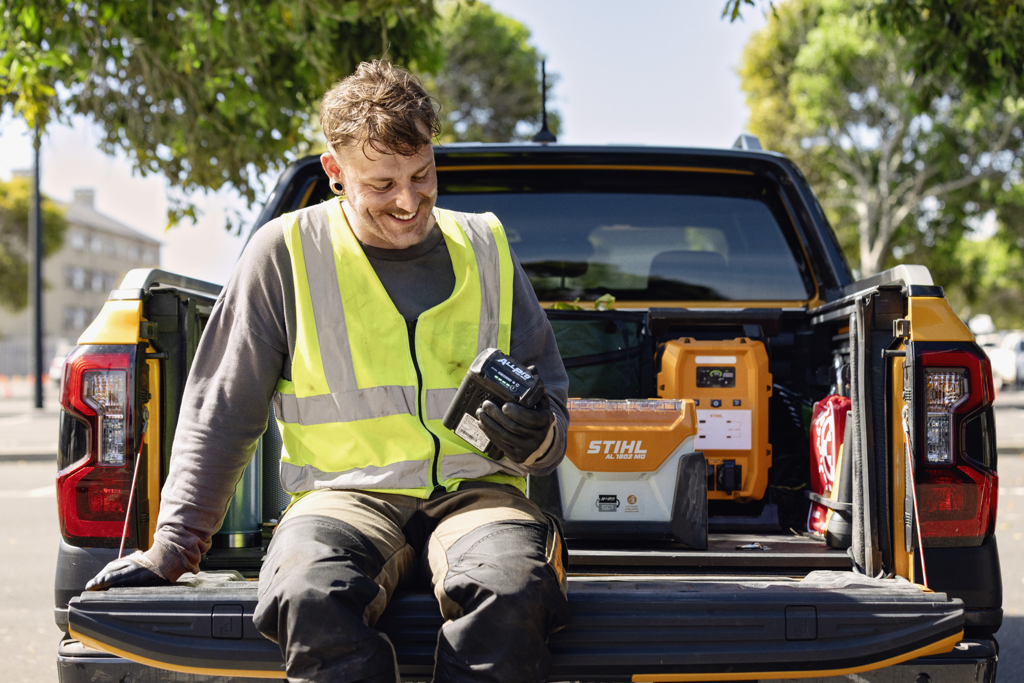 Mand, der sidder på bagsmækken af en pickup og tjekker opladningsniveauet på sit ALLPRO-batteri. I baggrunden ses STIHL AL 1802 MO hurtigladeren og den mobile strømforsyning PS 3000