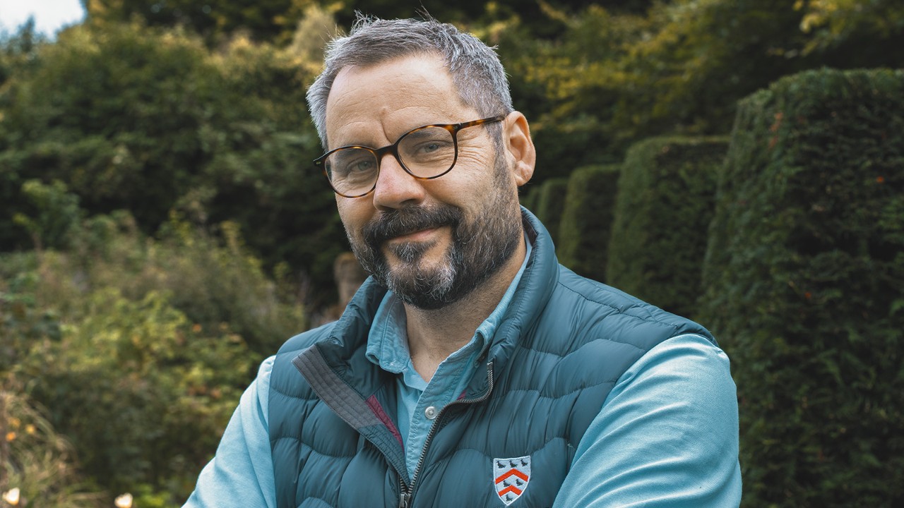 Portræt af lederen af Gardens and Grounds på Worcester College stående udendørs i en anlagt have, iført professionelt arbejdstøj og briller.