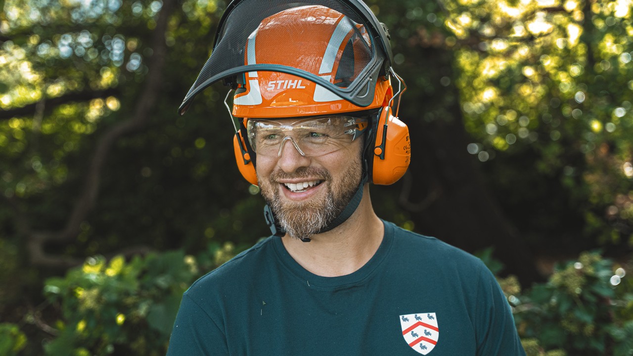 Portræt af en arborist fra Worcester College iført beskyttende hovedbeklædning og arbejdstøj, stående udendørs i et skovområde.