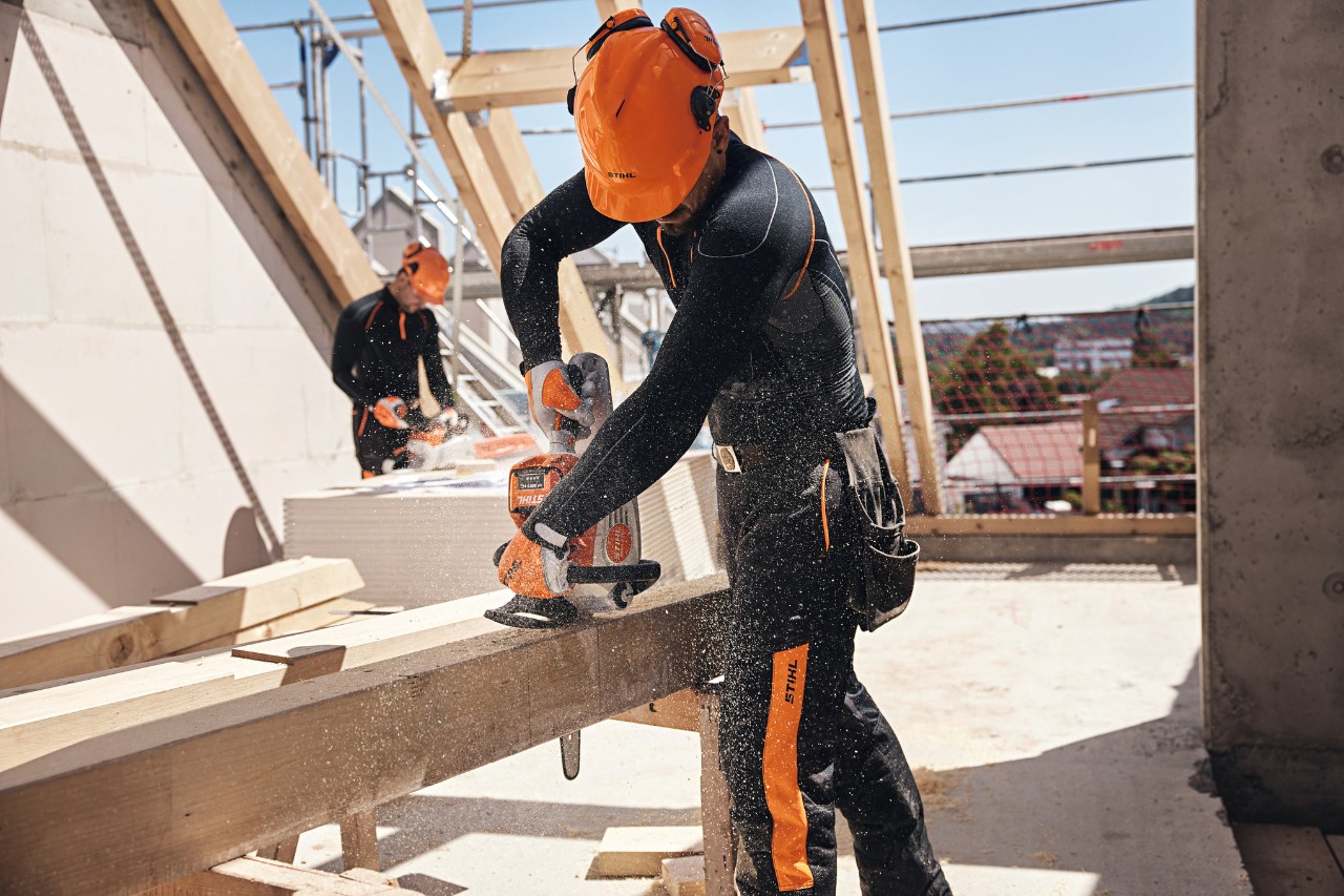 En STIHL-professionel på et loft saver en træbjælke med en batteridrevet motorsav, mens en anden arbejder bruger en batteridrevet motorsav i baggrunden