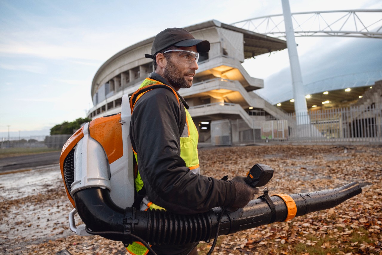 Sent på aftenen er en person i gang med at rydde blade og affald fra et stadion ved hjælp af den batteridrevne løvblæser ALLPRO STIHL BRA 600.